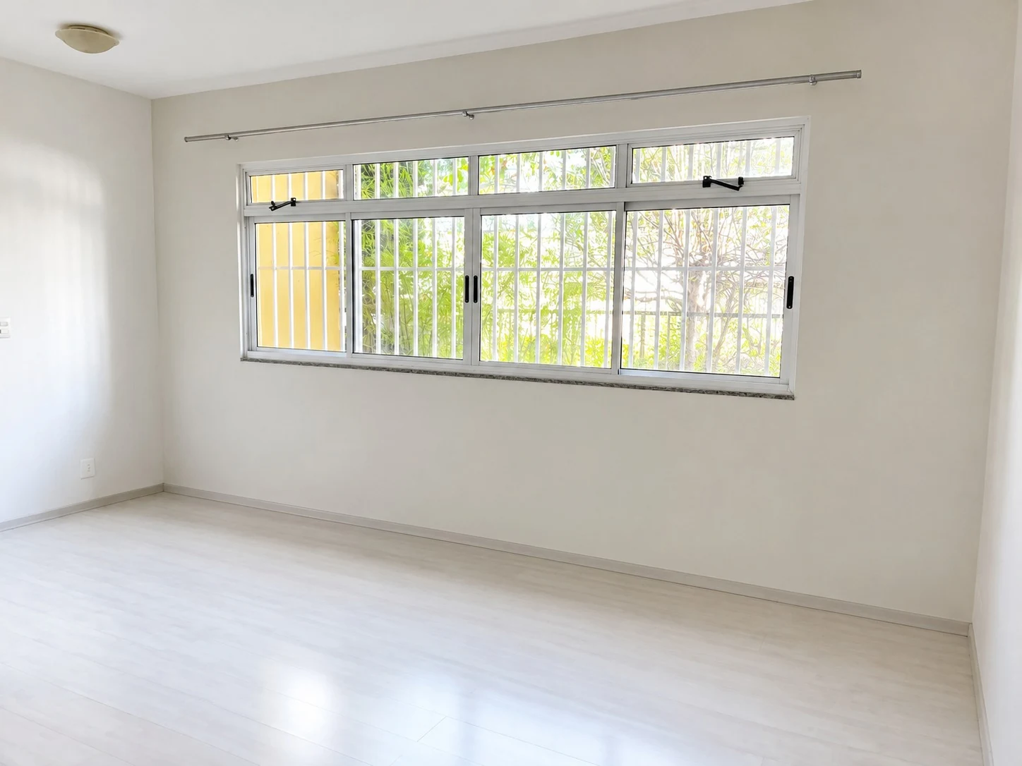 Sala clara e vazia, com janela ampla, paredes neutras e piso em padrão amadeirado.